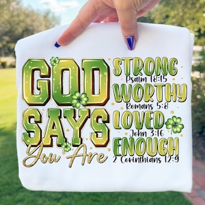 May include: White t-shirt with the text "GOD SAYS You Are STRONG WORTHY LOVED ENOUGH" in green and gold lettering. The design includes shamrock accents and Bible verses.