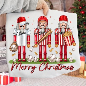 May include: A white pillow with three baseball-themed nutcracker figures in red and white uniforms, holding a baseball glove, bat, and popcorn. The words "Merry Christmas" are written in red script across the bottom. Baseball and peanut graphics are scattered around the figures.