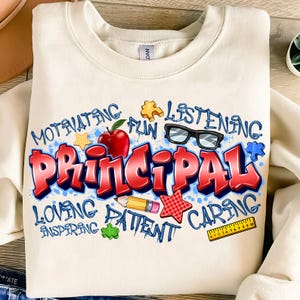 May include: Cream-colored sweatshirt with a colorful graphic design. The word "PRINCIPAL" is in large, red, graffiti-style letters. Surrounding the word are other words like "MOTIVATING", "LISTENING", and "CARING", along with various school-themed illustrations.