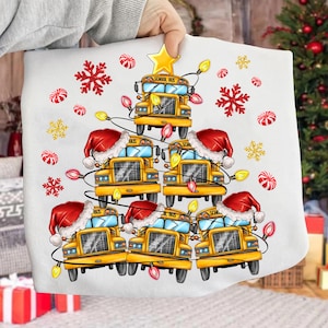 Op de afbeelding: Wit kussen met een kerstboomontwerp gemaakt van gele schoolbussen, elk met een kerstmuts. De boom is versierd met lichtslingers, sneeuwvlokken en zuurstokaccenten. Een gouden ster staat bovenaan.