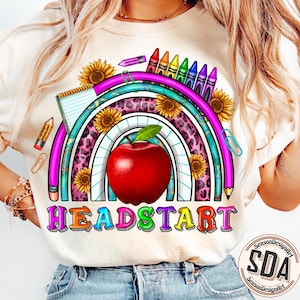 May include: A white t-shirt with a rainbow design featuring a red apple, sunflowers, and crayons. The text "HEADSTART" is printed in colorful letters below the rainbow.