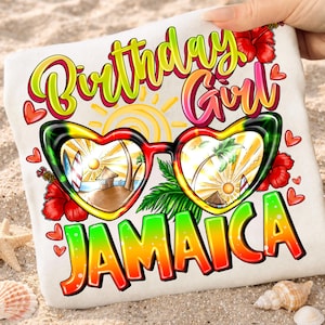 Può includere: Una maglietta bianca con un design grafico colorato. Il design presenta occhiali da sole a forma di cuore con una scena di spiaggia, le parole "Birthday Girl" e "Jamaica" in colori vivaci e fiori di ibisco rossi. La maglietta è tenuta da una mano.