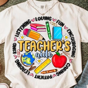 May include: White t-shirt featuring a colourful graphic for a teacher's aide. The design includes a pencil, books, apple, globe, and the words "Teacher's Aide" surrounded by words like "Loving" and "Kind".