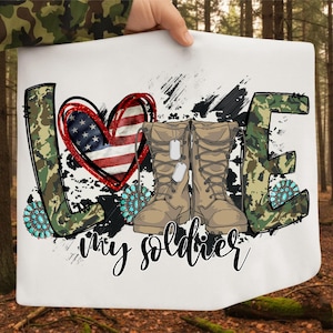 以下が含まれることがあります： 白い正方形の布に、迷彩柄の「LOVE」の文字とターコイズのアクセント。アメリカ国旗のデザインのハートと、ドッグタグ付きのコンバットブーツが特徴です。「my soldier」の文字が筆記体で書かれています。