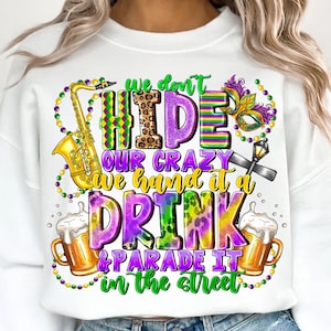 Puede incluir: Sudadera blanca con un colorido diseño de Mardi Gras. El diseño incluye un saxofón, jarras de cerveza, una máscara y el texto: "We don't hide our crazy, we hand it a drink & parade it in the street."