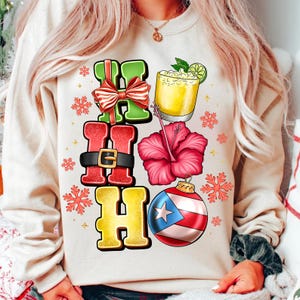 Puede incluir: Una sudadera de color crema con un diseño festivo. El diseño incluye las palabras "HO HO" en letras de bloque verdes, rojas y doradas, un cóctel, una flor de hibisco y un adorno con la bandera de Puerto Rico. Los acentos de copo de nieve completan el tema navideño.