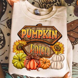 Könnte beinhalten: Cremefarbenes Sweatshirt mit einem herbstlichen Motiv. Das Design zeigt ein Holzschild mit der Aufschrift "Pumpkin Patch" in Orange und Gelb, Sonnenblumen, Kürbisse und eine braune Schleife.