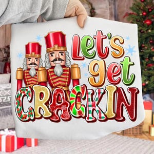 May include: White pillow with a festive design featuring two toy nutcracker soldiers in red and gold uniforms. The text "Let's Get Crackin" is displayed in colorful letters with snowflake accents, perfect for the holiday season.