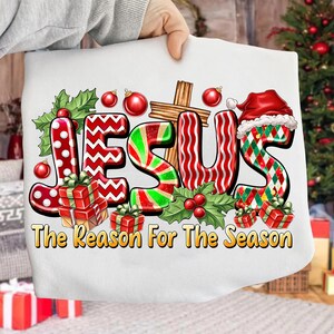 May include: White Christmas-themed graphic featuring the word "JESUS" in festive colors and patterns, including red, green, and white. The design incorporates a cross, holly, ornaments, and gift boxes, with the text "The Reason For The Season".