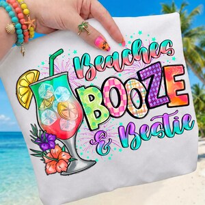 以下が含まれることがあります： カラフルなカクテルのグラフィックと「Beaches, Booze & Bestie」の文字がプリントされた白いTシャツ。カクテルは氷、レモンスライス、花が入った背の高いグラスに入っています。テキストは様々な色で表示されています。