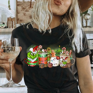 May include: Black t-shirt featuring a festive design with mugs decorated for Christmas. The mugs are adorned with candy canes, gingerbread men, and Grinch characters. The shirt also has the text "Alexa, Pour More Wine".
