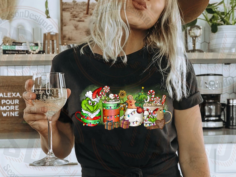 May include: Black t-shirt featuring a festive design with mugs decorated for Christmas. The mugs are adorned with candy canes, gingerbread men, and Grinch characters. The shirt also has the text "Alexa, Pour More Wine".