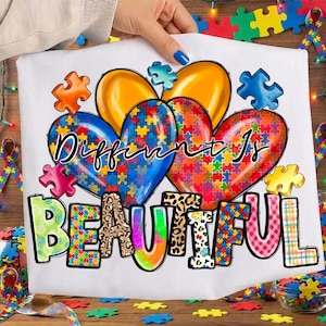 May include: White t-shirt with the words "Different Is Beautiful" in colorful, playful lettering. The design features puzzle piece hearts in blue, orange, and red, with puzzle pieces scattered around the shirt.