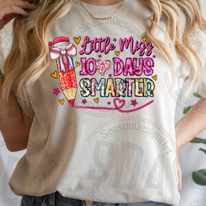 Op de afbeelding: Cr&egrave;mekleurig T-shirt met de tekst "Little Miss 100 Days Smarter" in roze en blauw. Een grote potlood met een roze strik en hartjes staat links. Het ontwerp bevat hartjes en sterren.