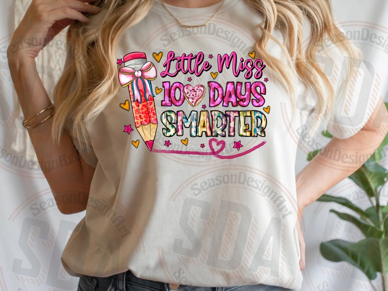 Op de afbeelding: Cr&egrave;mekleurig T-shirt met de tekst "Little Miss 100 Days Smarter" in roze en blauw. Een grote potlood met een roze strik en hartjes staat links. Het ontwerp bevat hartjes en sterren.