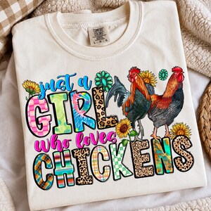 May include: A cream-colored t-shirt featuring a graphic print with two roosters, sunflowers, and turquoise accents. The text reads "just a GIRL who loves CHICKENS" in colorful, patterned letters including leopard print, plaid, and tie-dye designs.