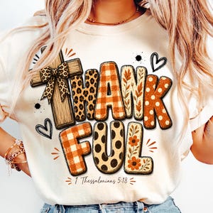 May include: A cream-colored t-shirt featuring the word THANKFUL in large, multi-patterned letters including orange plaid, leopard print, and floral designs. A wooden cross with a leopard print bow sits to the left, with the text 1 Thessalonians 5:18 below.