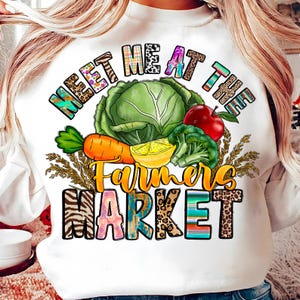 May include: White sweatshirt with a colorful graphic that reads "MEET ME AT THE FARMERS MARKET." The design features a cabbage, carrot, apple, broccoli, and lemon. The text and images have various patterns and textures.