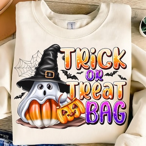 May include: Cream-colored sweatshirt featuring a Halloween-themed graphic. The design includes a ghost wearing a witch hat, a candy corn body, and a jack-o'-lantern bag. The words "Trick or Treat Bag" are displayed in orange and purple.