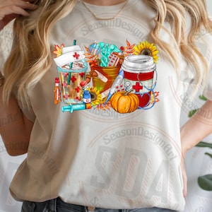 May include: A white t-shirt with a colorful graphic design featuring a coffee cup, a pumpkin, a first aid kit, and a syringe. The design includes fall-themed elements like sunflowers and leaves.