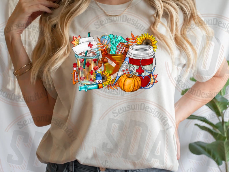 May include: A white t-shirt with a colorful graphic design featuring a coffee cup, a pumpkin, a first aid kit, and a syringe. The design includes fall-themed elements like sunflowers and leaves.