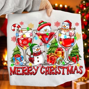 以下が含まれることがあります： クリスマスをテーマにした白い生地で、赤い液体が入った3つのワイングラスが描かれています。各グラスには雪だるまが入っています。グラスはストリングライト、雪の結晶、キャンディケインで飾られています。「MERRY CHRISTMAS」の文字は赤色です。