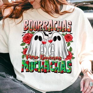 May include: White sweatshirt featuring a graphic of two ghost figures holding wine glasses. The text "BOORRACHAS Pero Buenas MUCHACHAS" is displayed above and below the ghosts, with red roses and bats as decorative elements.