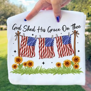 May include: White sweatshirt with the text "God Shed His Grace On Thee." The design features three American flags hanging on a clothesline, sunflowers, and green grass. The flags have red and white stripes and blue star fields.