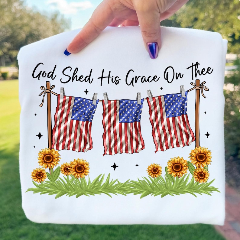 May include: White sweatshirt with the text "God Shed His Grace On Thee." The design features three American flags hanging on a clothesline, sunflowers, and green grass. The flags have red and white stripes and blue star fields.
