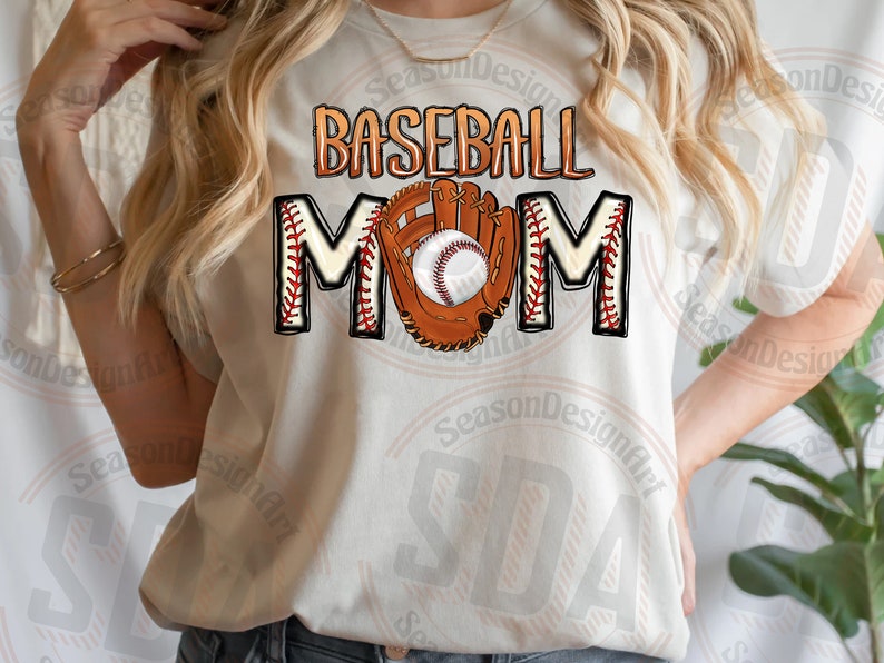 K&ouml;nnte beinhalten: Wei&szlig;es T-Shirt mit dem Text "Baseball Mom" in braunen und roten Buchstaben. Ein Baseballhandschuh mit einem Baseball darin befindet sich in der Mitte des Textes.