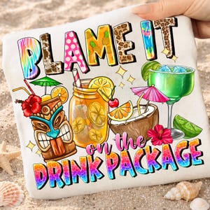 Könnte beinhalten: Ein weißes Strandtuch mit dem Text "BLAME IT ON THE DRINK PACKAGE" in bunten, stilisierten Buchstaben. Das Tuch zeigt Illustrationen von tropischen Getränken, darunter eine Tiki-Tasse, ein Einmachglas, eine Kokosnuss und ein Margarita-Glas.