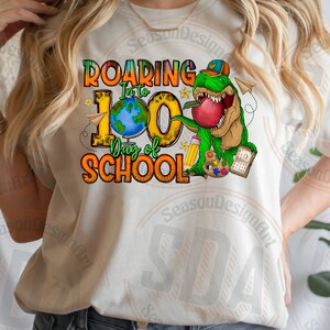 Op de afbeelding: Een wit T-shirt met een grafisch ontwerp van een groene dinosaurus die een appel vasthoudt. De tekst op het shirt luidt "Roaring into 100 Days of School".