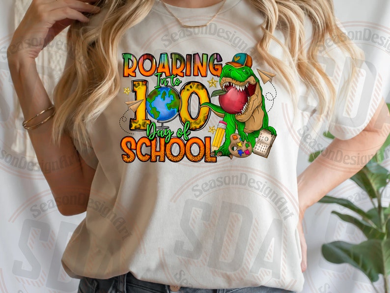 Op de afbeelding: Een wit T-shirt met een grafisch ontwerp van een groene dinosaurus die een appel vasthoudt. De tekst op het shirt luidt "Roaring into 100 Days of School".