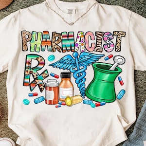 May include: White t-shirt with a colorful "PHARMACIST" graphic. The design includes a caduceus, mortar and pestle, prescription symbol, and scattered pills. The letters have animal print, stripes, and floral patterns.