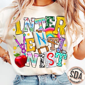 May include: Cream-colored sweatshirt with the word "INTERNIST" in colorful, whimsical lettering. The design includes a rainbow, apple, paint easel, and other school-themed graphics. The shirt has a crew neck and long sleeves.