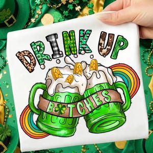 May include: White t-shirt with a St. Patrick's Day design. The shirt features two green beer mugs with overflowing foam, gold coins, and a rainbow. The words "DRINK UP B*TCHES" are printed on the shirt in various fonts and patterns.