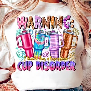 May include: White sweatshirt with the text "WARNING: I have an obsessive CUP DISORDER." The design features four colorful tumblers with various patterns, including tie-dye and leopard print. The word "WARNING" is in a mix of patterns.