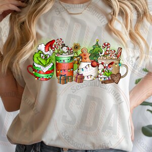 May include: A cream-colored t-shirt featuring a festive Christmas design. The design includes several mugs decorated with holiday characters like the Grinch, a snowman, and gingerbread men, along with candy canes and Christmas lights.