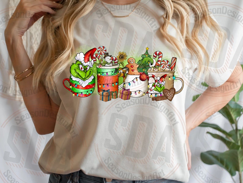 May include: A cream-colored t-shirt featuring a festive Christmas design. The design includes several mugs decorated with holiday characters like the Grinch, a snowman, and gingerbread men, along with candy canes and Christmas lights.