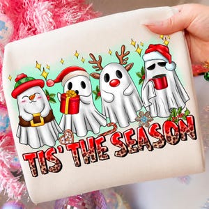 Könnte beinhalten: Ein cremefarbenes Sweatshirt mit einer Grafik von vier weißen Geistern, die für die Feiertage gekleidet sind. Einer trägt eine karierte Mütze, einer hält ein Geschenk, einer hat Rentiergeweihe und einer trägt eine Weihnachtsmütze. Text TIS' THE SEASON.