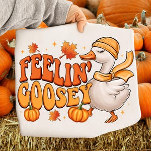 Könnte beinhalten: Ein weißes Sweatshirt mit einer Grafik einer Gans, die eine orangefarbene Mütze und einen Schal trägt. Der Text "FEELIN' GOOSEY" ist in großen orangefarbenen Buchstaben gedruckt, ergänzt durch Ahornblätter, Kürbisse und Sternakzente.