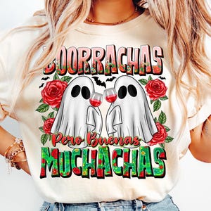 May include: Cream-colored t-shirt with a graphic featuring two ghost figures holding wine glasses, surrounded by red roses and green leaves. The shirt has the words "Borrachas Pero Buenas Muchachas" in a colorful, decorative font.
