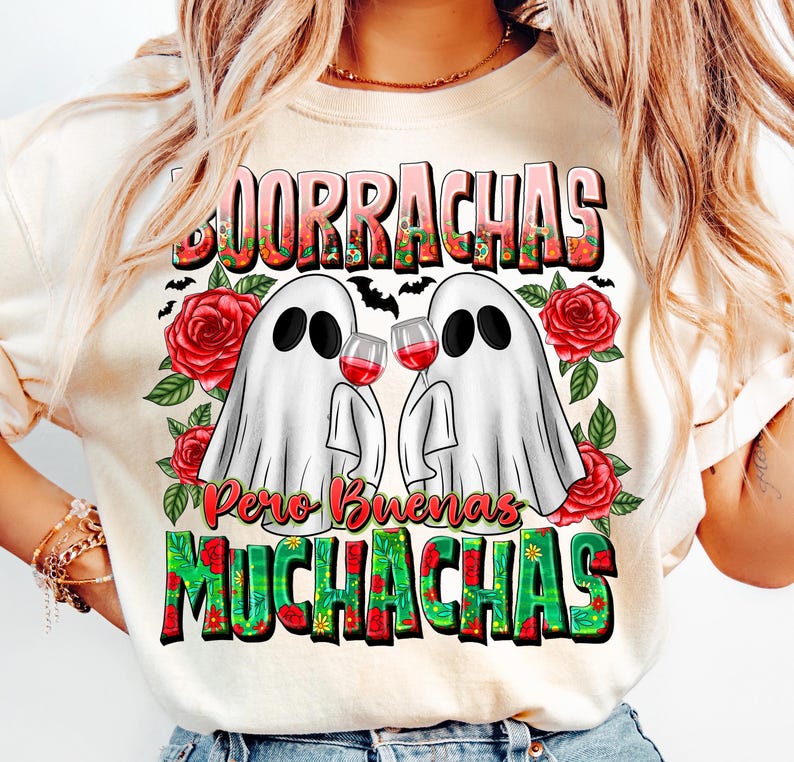 May include: Cream-colored t-shirt with a graphic featuring two ghost figures holding wine glasses, surrounded by red roses and green leaves. The shirt has the words "Borrachas Pero Buenas Muchachas" in a colorful, decorative font.