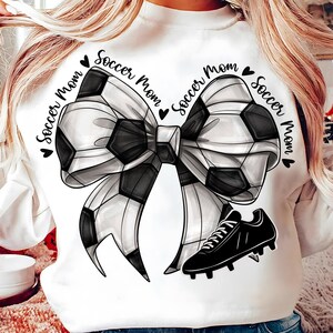 May include: White sweatshirt featuring a soccer ball bow design with the words "Soccer Mom" and a black soccer cleat. The bow is black and white, and the text is in a handwritten style.