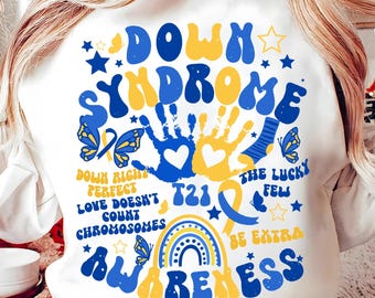 Pode incluir: Camisola branca com um design gráfico azul e amarelo que diz "Down Syndrome Awareness". O gráfico inclui impressões de mãos, um arco-íris, borboletas e o texto "Down Right Perfect", "T21", "The Lucky Few", e "Be Extra".