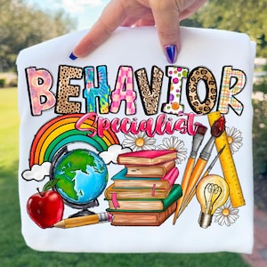 Può includere: Felpa bianca con la scritta "BEHAVIOR Specialist" in un design colorato e fantasia. Il disegno include un arcobaleno, un mappamondo, libri, una mela, pennelli e una lampadina, per gli educatori.