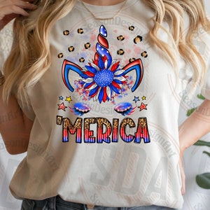 Może przedstawiać: Biały T-shirt z wzorem jednorożca w kolorze czerwonym, białym i niebieskim. Jednorożec ma słonecznik jako grzywę, a napis 'MERICA' w czerwonych, białych i niebieskich literach z tłem w panterkę.
