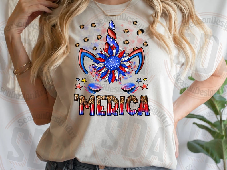 Może przedstawiać: Biały T-shirt z wzorem jednorożca w kolorze czerwonym, białym i niebieskim. Jednorożec ma słonecznik jako grzywę, a napis 'MERICA' w czerwonych, białych i niebieskich literach z tłem w panterkę.