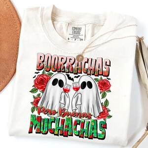 May include: Cream-colored t-shirt with a graphic featuring two ghost figures holding wine glasses, surrounded by red roses and the text "BOORRACHAS Pero Buenas MUCHACHAS". The shirt has a Comfort Colors label and a gold necklace.