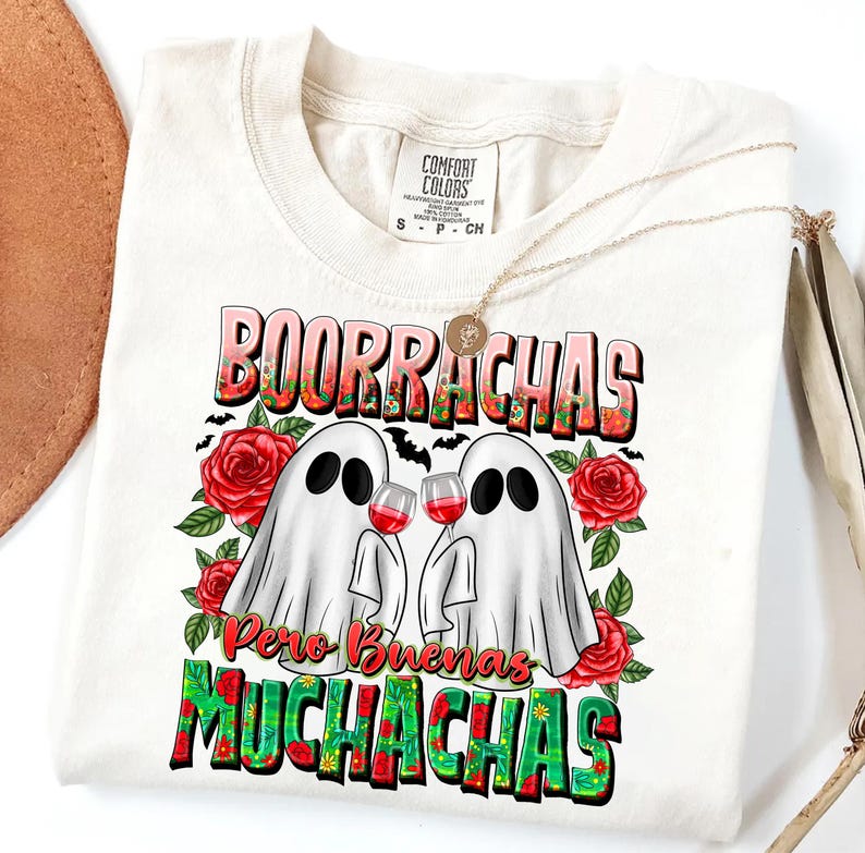 May include: Cream-colored t-shirt with a graphic featuring two ghost figures holding wine glasses, surrounded by red roses and the text "BOORRACHAS Pero Buenas MUCHACHAS". The shirt has a Comfort Colors label and a gold necklace.
